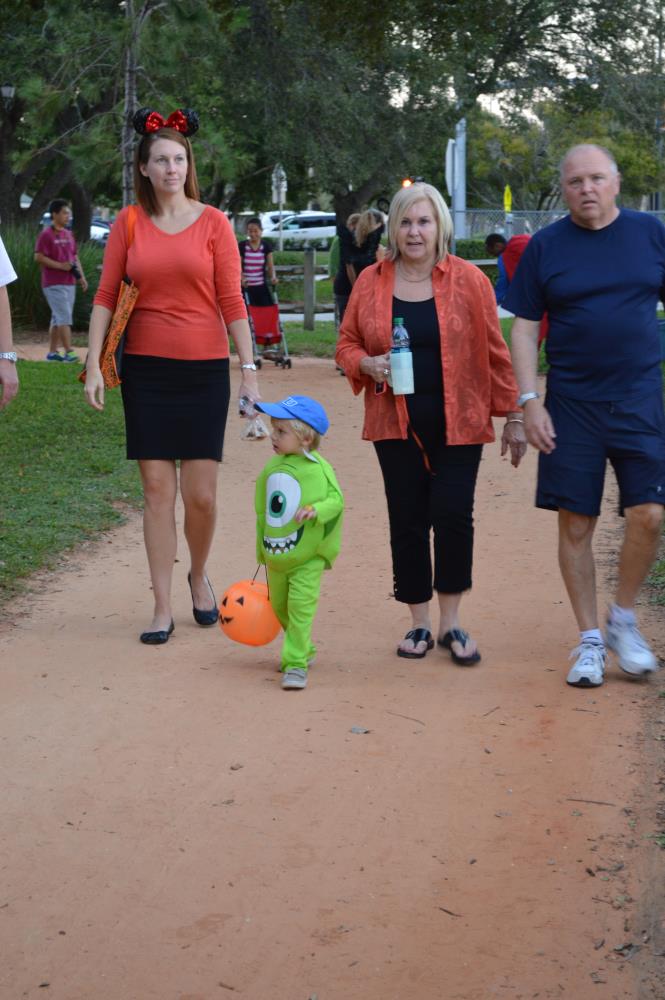 Kid dressed as Mike Wazowski carrying orange jack-o-lantern walks with adults